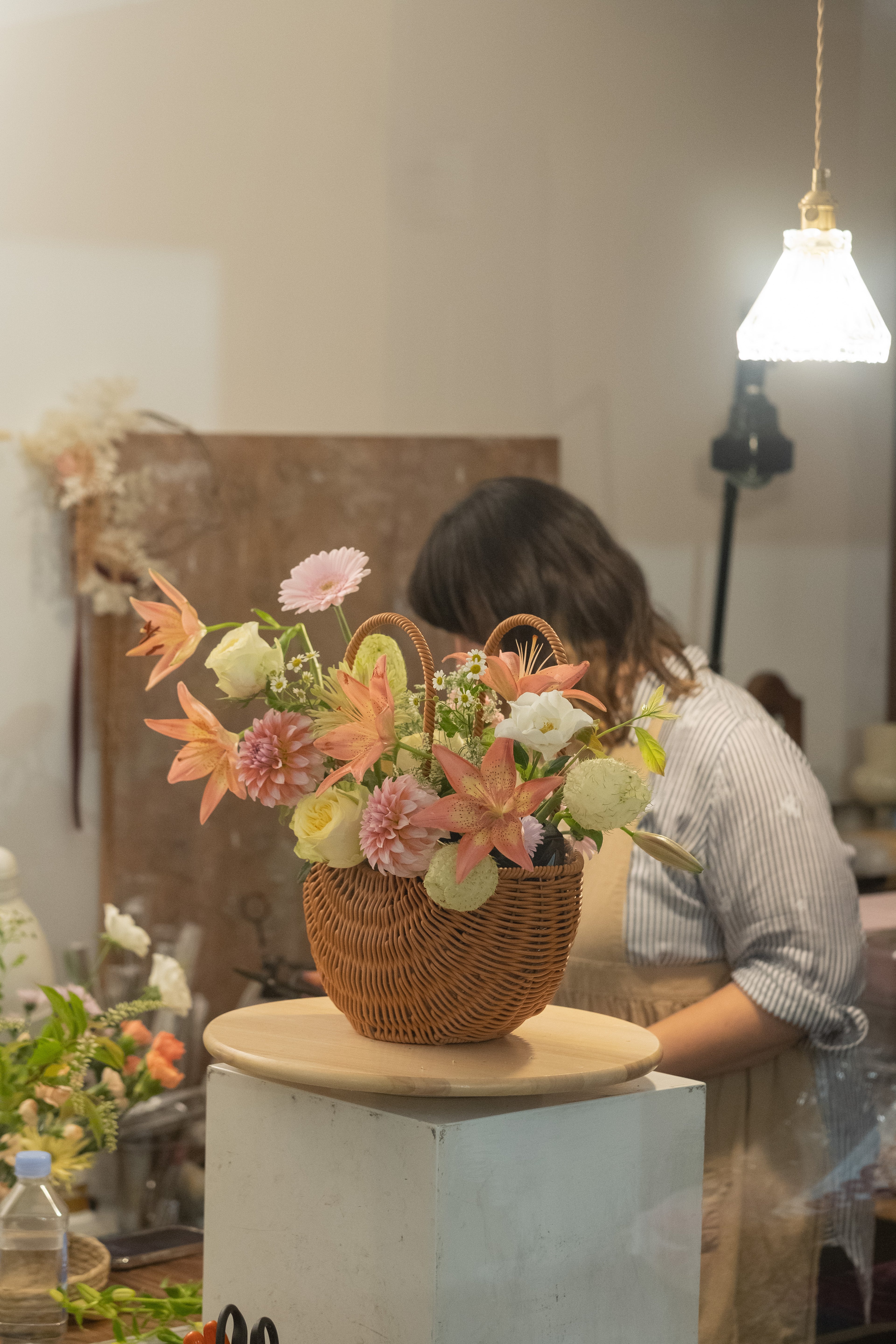 Floral arrangement in a woven basket on a wooden stand with a blurred background of a person and flowers.